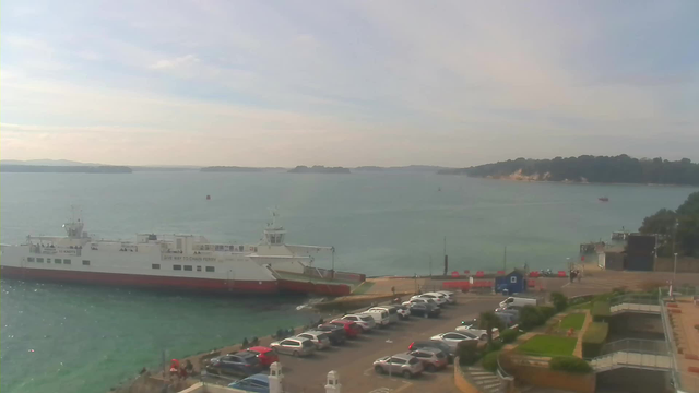 A view of a calm body of water with a large white ferry docked at a pier on the left. The ferry has red stripes and a sign indicating "10 KNOTS" and "GIVE WAY TO CHAIN FERRY." In the foreground, there is a parking lot with several cars parked, and some people are visible near the waterfront. The background features a distant shoreline with trees, and gentle waves in the water under a partly cloudy sky.