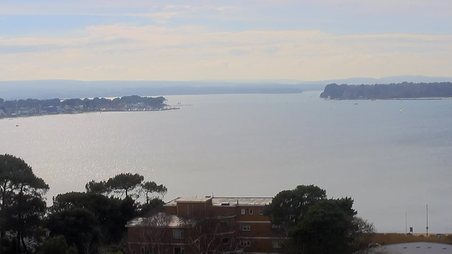 A serene waterfront scene with calm water reflecting sunlight, framed by distant hills and a cloudy sky. In the foreground, there is a low building surrounded by trees. To the left, a shoreline with small boats is visible, while the water stretches towards the horizon, where it meets the sky. The overall atmosphere is tranquil and natural.