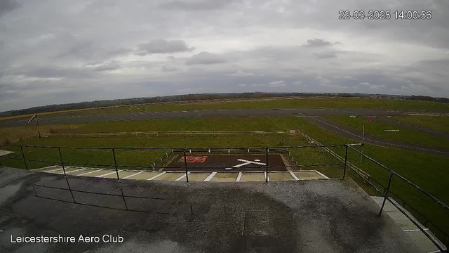 A view from a webcam showing an open-air area at the Leicestershire Aero Club. In the foreground, there is a flat rooftop with a railing. Below, there is a grassy area with a white X marking a landing spot, and a red ground marking nearby. To the left, a windsock is visible, indicating the wind direction. The background features expansive green fields and a cloudy sky, suggesting an overcast day.