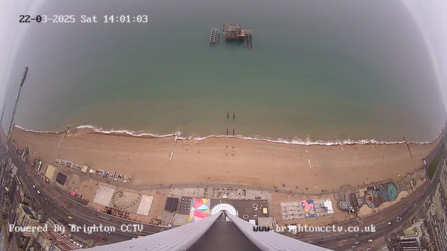 Aerial view of a sandy beach with light brown sand and gentle waves lapping at the shore. In the foreground, a pier structure extends into the ocean, partially submerged. There are several beachgoers walking along the shore, and a few silhouettes in the water. The beach is lined with colorful beach huts and attractions, with a prominent circular structure and a multicolored patterned area visible. The sky is overcast and gray. A timestamp in the top left corner indicates the date and time as March 22, 2025, at 14:01:03.
