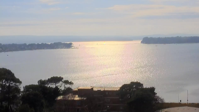 A serene view of a large body of water reflecting sunlight, creating a shimmering effect on its surface. In the background, a shoreline with trees and buildings visible, as well as distant hills and small boats dotting the water. The sky is light with soft clouds, and the overall scene conveys a peaceful atmosphere.