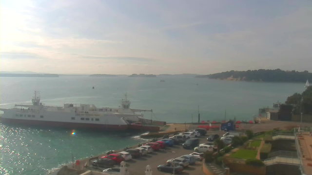 A view of a harbor with a large white ferry docked at a terminal. The water is shimmering in shades of blue and green. There are several boats in the distance and a line of parked vehicles along the shore, with a mix of colors including red, white, and blue. In the background, there are low hills and trees lining the shore. The sky is partly cloudy, and the scene appears calm and serene.