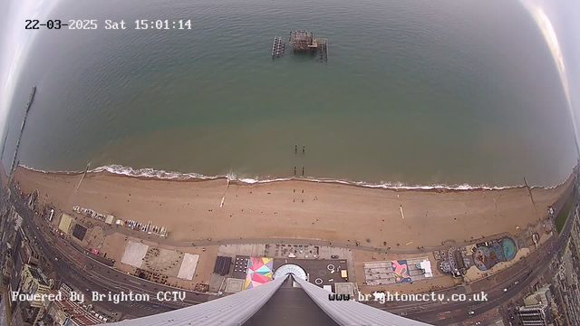 Aerial view of a beach with varying shades of sand and shallow waves. In the distance, a broken pier emerges from the water, while several people can be seen walking along the shore. Along the beach, there are colorful structures and a carnival area with various attractions. The bottom of the image shows a wide walkway and buildings lining the seafront. The sky is mostly clear, indicating daylight.