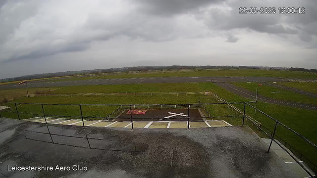 A wide view of a runway at the Leicestershire Aero Club, featuring a grassy field with marked boundaries. The scene is overcast, with dark clouds overhead. In the foreground, there is a white "X" on the ground indicating a location, and a partially visible building structure with railings. A gravel area is present beside the runway, and distant structures are visible in the background. The ground appears wet, suggesting recent rain.