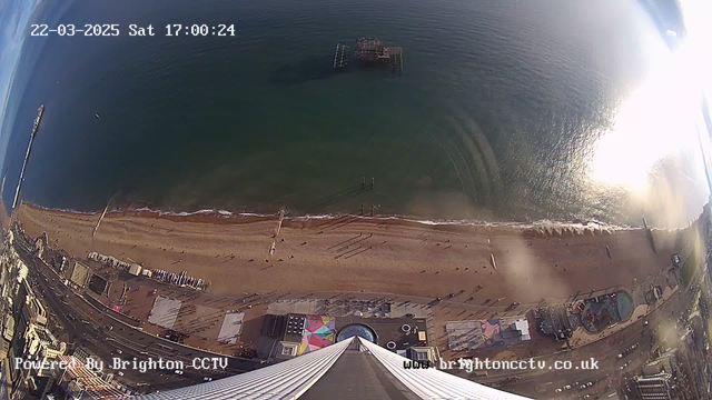 Aerial view of a beach with light brown sand and a gentle shoreline meeting the blue sea. In the distance, an old pier extends into the water, visible as a metal structure. The beach is dotted with people and various structures, including colorful beach huts and a circular swimming pool, probably for leisure activities. The ocean reflects sunlight, creating sparkling patterns on the surface. The scene is framed by buildings along the edge of the beach, showing the contrast between urban and natural environments. The image timestamp shows it is Saturday, March 22, 2025, at 17:00:24.
