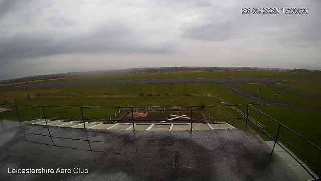 A view from a webcam showing the Leicestershire Aero Club. The image features a large green field with a runway made of asphalt. In the foreground, there is a railing and some steps leading down from the platform. The sky is overcast with gray clouds, and there is a slight visibility of the surrounding landscape in the distance. The ground is wet, likely indicating recent rain.