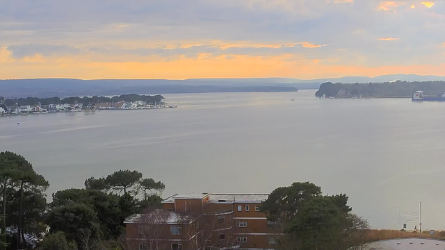A serene view of a calm body of water reflecting soft hues of orange and gray from the sky at sunset. In the foreground, there are low trees, followed by a building with multiple windows. Beyond the building, several boats are visible on the water near a shoreline lined with houses and docks. The distant landscape features rolling hills under a cloudy sky.