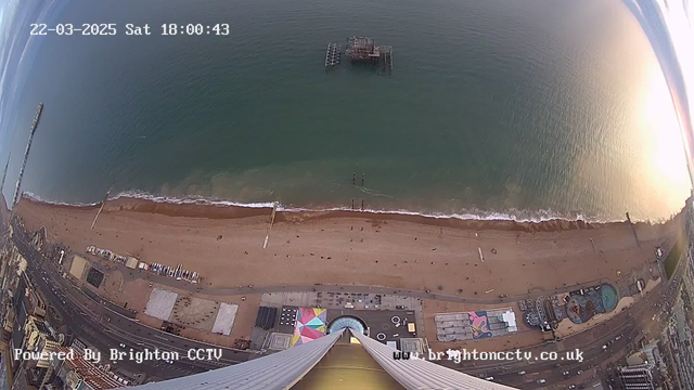 A high-angle view of a beachfront scene, taken from a webcam. The image shows a sandy beach with a few people scattered along the shore, and gentle waves lapping at the water's edge. To the left, there is a wooden pier extending into the sea, and further out, a partially submerged structure can be seen. The beach is lined with colorful beach huts and a fairground area with visible rides and attractions. Above, the sky is mostly clear with some clouds, and the time display indicates it is Saturday, March 22, 2025, at 18:00:43.