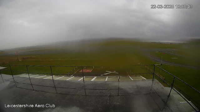 A view from a webcam at the Leicestershire Aero Club showing a rainy and overcast scene. The foreground includes a railing and a flat surface with white lines. In the background, there is an open field and a runway, partially visible through the haze and rain. The date and time are displayed at the top right corner, indicating it is the 22nd of March, 2025, at 6:00 PM.