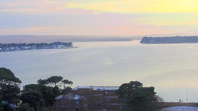 A serene landscape featuring a calm waterway reflecting pastel colors of a sunrise or sunset. In the foreground, there are several buildings partially obscured by trees. The distant shoreline features a marina with boats and similar buildings against rolling hills in the background. The overall atmosphere is tranquil, with soft colors blending in the sky.