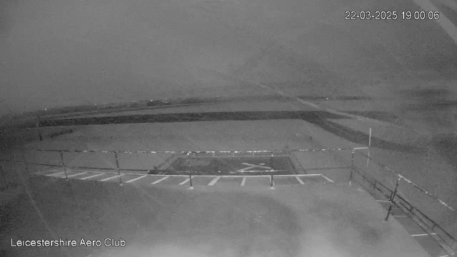 A grayscale image from a webcam captures an empty outdoor space at dusk. In the foreground, there is a fenced area with several empty parking spaces marked by white lines. The ground is flat and slightly visible in the dim light. In the background, there are blurred outlines of fields and a horizon, suggesting a rural setting. The timestamp at the top right indicates the date and time as March 22, 2025, at 19:00:06.