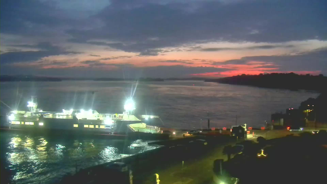 A ferry is docked at a pier, illuminated by bright lights. The water reflects these lights, creating a shimmering effect. In the background, the sky is transitioning to twilight, showcasing shades of blue, purple, and pink as the sun sets. Silhouettes of distant land can be seen on the horizon, and various vehicles are parked near the dock.