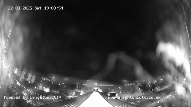 A black and white webcam image showing a city landscape at night. The bottom of the image features a tall structure with a pointed roof, leading the eye upwards toward a dark sky. The area beneath the structure exhibits streets and buildings, with some lights visible indicating vehicles or street lamps. There is a timestamp in the top left corner reading "22-03-2025 Sat 19:00:54," and a watermark at the bottom left stating "Powered By Brighton CCTV."