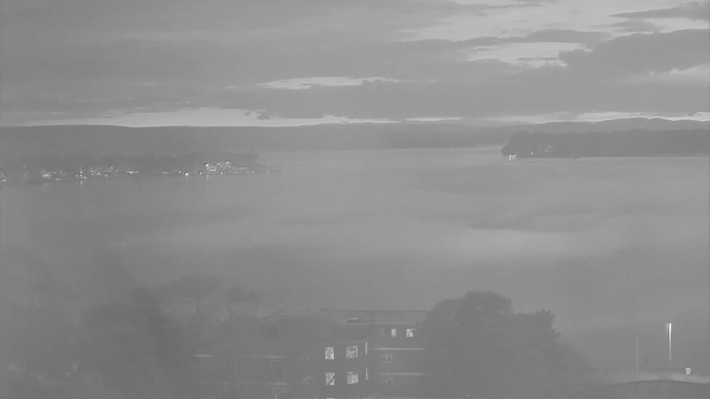 A scenic view of a body of water, surrounded by low hills and a cloudy sky. The image is in black and white, creating a calm and moody atmosphere. In the foreground, there are silhouettes of trees and a building with windows that are slightly illuminated. In the background, faint lights reflect off the water, suggesting distant structures along the shoreline. The sky above has varying shades, hinting at a sunset or sunrise in progress.