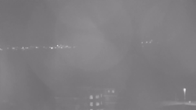A monochromatic image depicting a night scene with blurred outlines. In the foreground, a dark structure is visible, possibly a building with faint light reflections. In the background, there are scattered lights that suggest distant objects, such as streetlights or buildings, but they are obscured by a haze or fog, creating a soft focus effect. The overall atmosphere is dim and obscured, with no distinct features presented clearly.