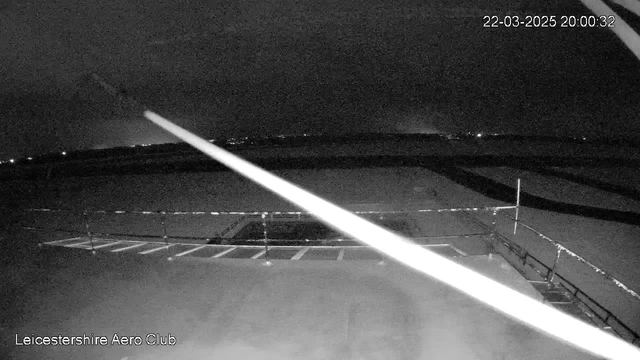A dimly lit scene of an airfield at night. In the foreground, there is a flat surface with marked parking lines partially visible. A white streak, possibly a light or reflection, runs diagonally across the image from the top left to the bottom right. In the background, faint lights can be seen in the distance, suggesting the presence of buildings or vehicles, while the sky is dark. The date and time are displayed in the top right corner.