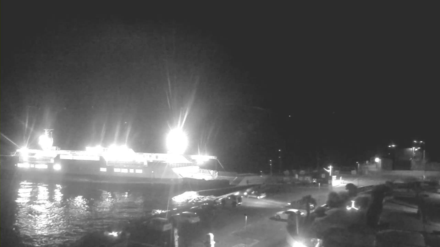 A brightly lit ferry is docked at a harbor at night, reflecting light on the water. The area surrounding the ferry includes various parked vehicles and dimly lit structures in the background. The scene is illuminated predominantly by the ferry's lights, creating a stark contrast against the dark surroundings.