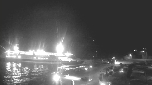 A nighttime scene at a harbor with a large ferry illuminated by bright lights. The ferry is docked at the edge of the water, with reflections visible on the surface. In the foreground, there are several parked cars and structures along the waterfront, while additional faint lights can be seen in the background, including buildings and streetlights. The overall atmosphere is dark, with the ferry being the main source of illumination.