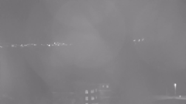 A gray-scale image showing a foggy or hazy view. Vague outlines of buildings are visible in the foreground, with some light points suggesting streetlights or distant structures in the background. The overall scene appears obscure due to reduced visibility.