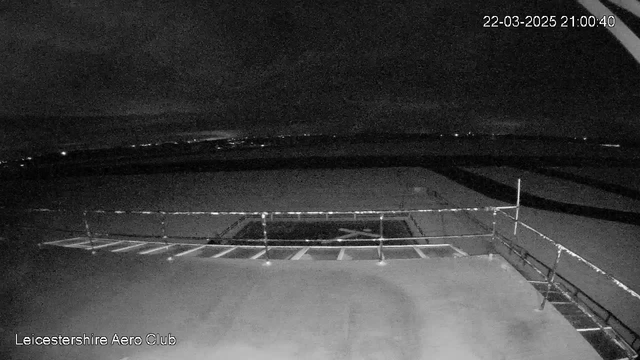 A nighttime image shows a flat rooftop with a railing surrounding one section. The background features a dark sky with scattered lights, indicating a distant urban area or horizon. The scene appears to be taken from a webcam at the Leicestershire Aero Club, showing minimal detail due to low lighting conditions.