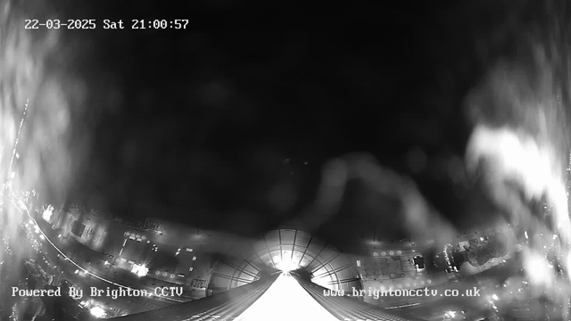 A webcam image showing a dark scene illuminated by faint light. The view appears to be from a high location looking downward along a pathway or roadway. There are blurred outlines of structures on either side, suggesting buildings or streetlights. The image has a timestamp at the top indicating the date and time. The bottom left corner includes text that reads "Powered By Brighton CCTV" and a website address in the lower right corner.