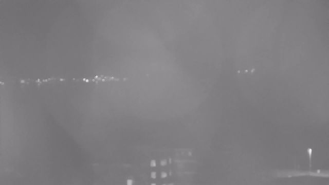 A monochrome image taken at night, displaying a cloudy or foggy scene. In the background, faint lights from distant buildings or streetlights create a hazy line. The foreground features an indistinct silhouette of structures, possibly buildings or trees, partially obscured by the atmospheric conditions.