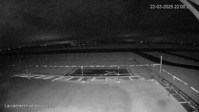 A dimly lit scene of an outdoor area, possibly a runway or airfield, observed from a high vantage point. The foreground features a flat surface with a white cross marking, surrounded by a metal railing. In the distance, there are faint lights indicating buildings or vehicles. The sky is mostly dark with some clouds, and the overall atmosphere is quiet and still, as it appears to be nighttime. The timestamp indicates the image was captured on March 22, 2025, at 10:00 PM.