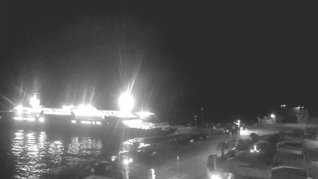A nighttime scene at a dock, featuring a large illuminated ferry on the water. The ferry has bright lights shining from its structure. In the foreground, the dock is partially visible with several vehicles parked. The water reflects the lights from the ferry, creating a glimmering effect. The background is dark, with some lights illuminating the dock area and nearby structures.