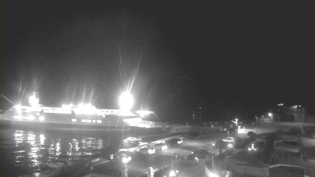 A nighttime scene at a harbor, featuring a large illuminated ferry docked at the edge of the water. The ferry is surrounded by several bright lights. In the foreground, there are various parked cars and a road leading toward the terminal area, which is also illuminated by street lights. The sky is dark, with minimal visibility of distant features.