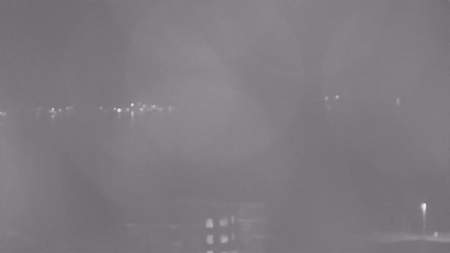 A hazy night scene captured by a webcam, showing faint lights reflected on a body of water. The image is predominantly gray, with soft circular shapes indicating water ripples and light sources. A few lights can be seen in the background, suggesting the presence of distant buildings or boats. The foreground is obscured by fog or mist, creating a subtle and indistinct appearance.