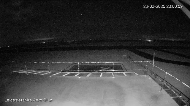 A dark scene captured by a webcam, showing a flat surface possibly a rooftop or landing area. There is a railing visible along the front edge. In the background, faint lights from distant structures or vehicles can be seen, and the sky is mostly dark with a hint of clouds. The timestamp in the corner indicates the date and time of the image.
