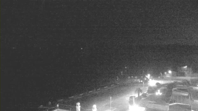A dark scene captured by a webcam, with minimal visibility. The foreground features a partially illuminated area suggesting a parking lot or open space with a few vehicles. In the background, faint lights from structures and possibly distant vehicles indicate activity. The overall image has low contrast due to the night setting, and the surrounding area appears shadowy and indistinct.