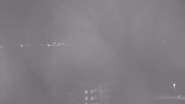 A blurred black-and-white image showing a dimly lit scene at night. There are scattered points of light in the background, likely streetlights or reflections, and a darker shape in the foreground resembling a building or surface. The overall visibility is low, creating a foggy or misty atmosphere.