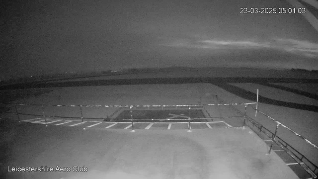 A dark scene captured from a webcam showing the exterior of Leicester Aero Club. The view includes an empty runway with markings and parked aircraft stands visible. A railing runs along the foreground, with a partially cloudy sky above. The image is in black and white, suggesting early morning or dusk hours. The timestamp in the corner indicates the date and time of the capture, which is March 23, 2025, at 5:01 AM.
