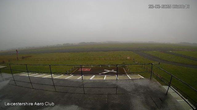 A view from a webcam at the Leicestershire Aero Club, showing a foggy scene. In the foreground, there is a railing and a concrete surface. Below the railing, a large "X" marking is visible on the ground, with a red rectangle beside it. The background features a grassy area and a runway that stretches into the distance, slightly obscured by fog. There are fence lines marking the edges of the field, and the sky appears grey and overcast. The time displayed is 06:00 on March 23, 2025.