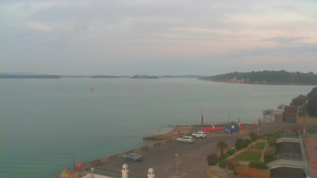 A serene view of a waterfront scene under a cloudy sky. The calm water stretches to the horizon, where small islands can be seen in the distance. A few boats can be spotted on the water. In the foreground, there is a parking area with several vehicles, including two cars parked side by side. A small red buoy floats in the water, and there are hints of green landscaping near the edge of the parking lot, as well as structures along the shoreline.