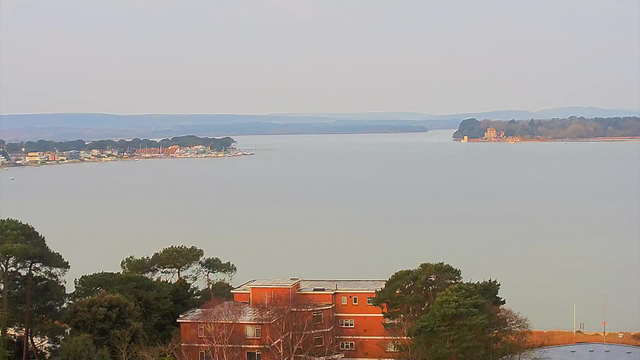 A panoramic view of a calm body of water, with a shoreline featuring buildings and trees. In the foreground, there is an orange brick building surrounded by greenery. To the left, several boats are visible near the distance, along with a row of colorful buildings lining the shore. On the right side, an island appears with a structure resembling a castle. The background consists of rolling hills under a subtle, overcast sky, creating a serene atmosphere.