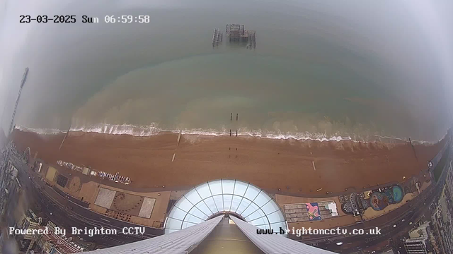 An aerial view of a beach scene captured from a webcam, taken early in the morning on March 23, 2025. The image shows a sandy beach with gentle waves lapping at the shore. In the water, a distant pier structure is visible, partially submerged. The foreground features a circular building structure, possibly a viewing platform, with a clear glass roof. To the right, there are hints of colorful beach attractions and facilities. The sky appears overcast, contributing to a moody atmosphere. The timestamp in the top left corner reads "06:59:58".