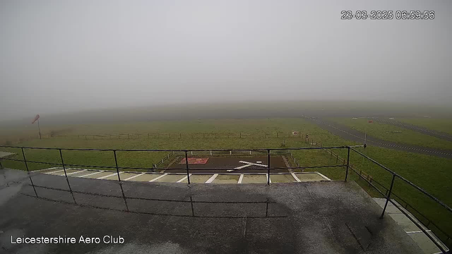 A foggy scene viewed from a raised platform at the Leicestershire Aero Club. The ground below is mostly grass, with a dark marked area resembling a runway or helipad featuring a white cross. There is a windsock on the left side, indicating light wind conditions. Visibility is low due to the thick fog, obscuring distant objects and making the surrounding environment appear gray and muted.