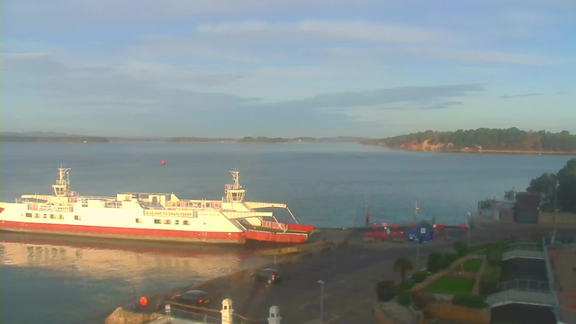 A ferry docked at a port is visible in the foreground, while calm water reflects the sky. In the background, there are hills and a shoreline lined with trees, with a faint mist hovering above the water. The scene is bathed in soft morning light, and a few vehicles are parked near the ferry terminal.