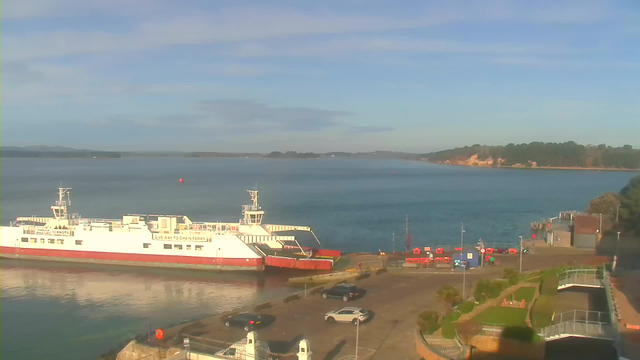 A large white ferry with red trim is docked at a port, which is located by a calm blue body of water. The ferry has a sign indicating a speed limit of 15 knots. In the background, there are low hills and a sandy shoreline under a clear blue sky. The dock area features several parked cars and a few orange safety buoys in the water. There are glimpses of greenery and walkways near the dock.