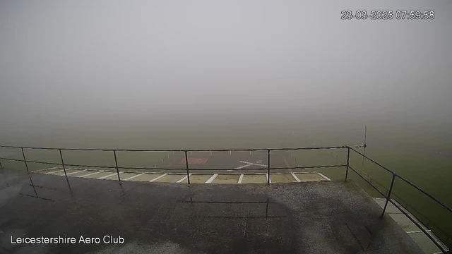 A view from a balcony overlooking a foggy landscape. The foreground features a metal railing slightly obstructing the view. Below, there is a flat surface with visible stripes, leading to a large, indistinct area of grass blurred by the fog. The background is entirely shrouded in thick mist, making it difficult to see any further details or objects. The image is timestamped March 23, 2025, at 07:59:58, and is associated with the Leicestershire Aero Club.