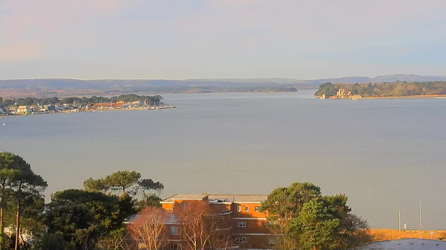 A panoramic view of a calm body of water, with a shoreline lined with buildings and trees. In the foreground, there is an arrangement of trees and buildings, while further back, the water stretches into the distance, leading to more buildings and greenery along the opposite shore. Rolling hills are visible in the background under a clear sky. The scene conveys a tranquil outdoor setting.
