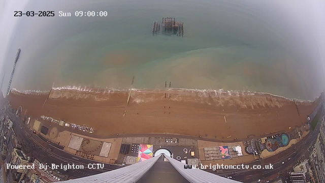 A high-angle view of a beach with brown sand and gentle waves lapping at the shore. In the background, a calm blue sea meets the horizon. There is a partially submerged pier in the water and a few small figures walking along the beach. Colorful beach huts and attractions are visible on the shoreline, with a large circular structure near the bottom of the image. The sky is overcast, suggesting a cloudy day. A date and time stamp is displayed in the upper left corner.