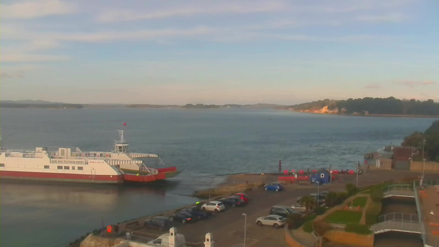 A large ferry is docked at a harbor with a colorful red and white pattern. The calm water surrounds the boat, reflecting the sky. In the background, there are hilly islands, and the coastline features trees and a light-colored cliff. In the foreground, a parking area is visible with several cars parked. There are also bright, round objects near the water and a few small structures along the shore. The sky is partly cloudy, creating a serene atmosphere.