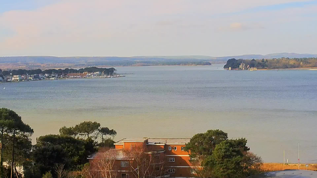 A wide view of a calm body of water, with land visible on both sides. On the left side, there’s a marina with several boats and buildings along the shore. In the foreground, a low building with a mix of windows and brown walls can be seen, surrounded by trees. In the background, rolling hills are visible under a clear sky. The sunlight creates a soft reflection on the water's surface, which features gentle waves.