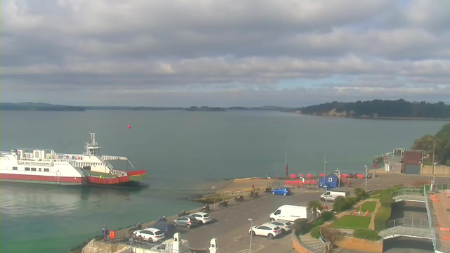 A view of a calm harbor with a ferry approaching from the left side. The ferry has a white hull with red accents and is partially docked. In the foreground, there are several parked cars. The parking area is adjacent to the water and has well-maintained green spaces. The sky is partly cloudy with patches of blue, and several boats can be seen in the distance.