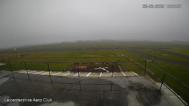 A view from a webcam at the Leicestershire Aero Club shows a foggy landscape. The foreground features a sloped railing, while the background reveals an airfield covered in grass with a runway and a helipad marked by white lines. Visibility is low due to fog, obscuring distant objects and making the scene appear muted and gray. A flag can be seen on a pole to the left, signaling the direction of the wind. The date and time are displayed in the upper right corner of the image.