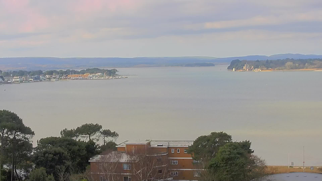 A serene view of a body of water under a cloudy sky. The foreground features a building with a flat roof and several trees. In the distance, a shoreline stretches along the water, dotted with small boats and houses. The landscape includes hills that rise gently in the far background. The overall atmosphere is calm and peaceful.