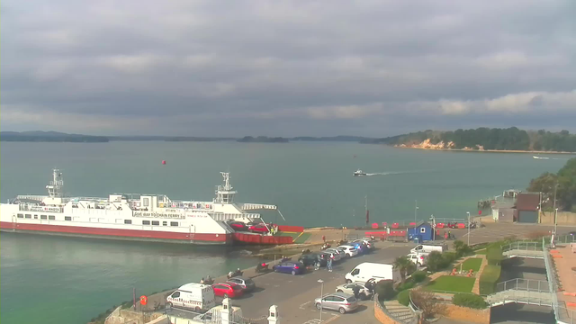 A view of a harbor with a large ferry docked at the pier, displaying red and white colors. Several cars and people are visible along the waterfront. In the background, a small boat is navigating through the water. The sky is partly cloudy, and a shoreline with greenery can be seen in the distance. The scene captures a tranquil marine atmosphere.